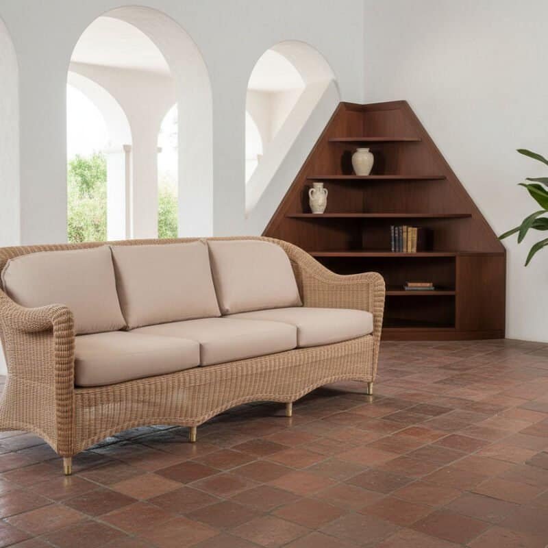 Divano in vimini beige in soggiorno luminoso con libreria in legno e pianta verde.