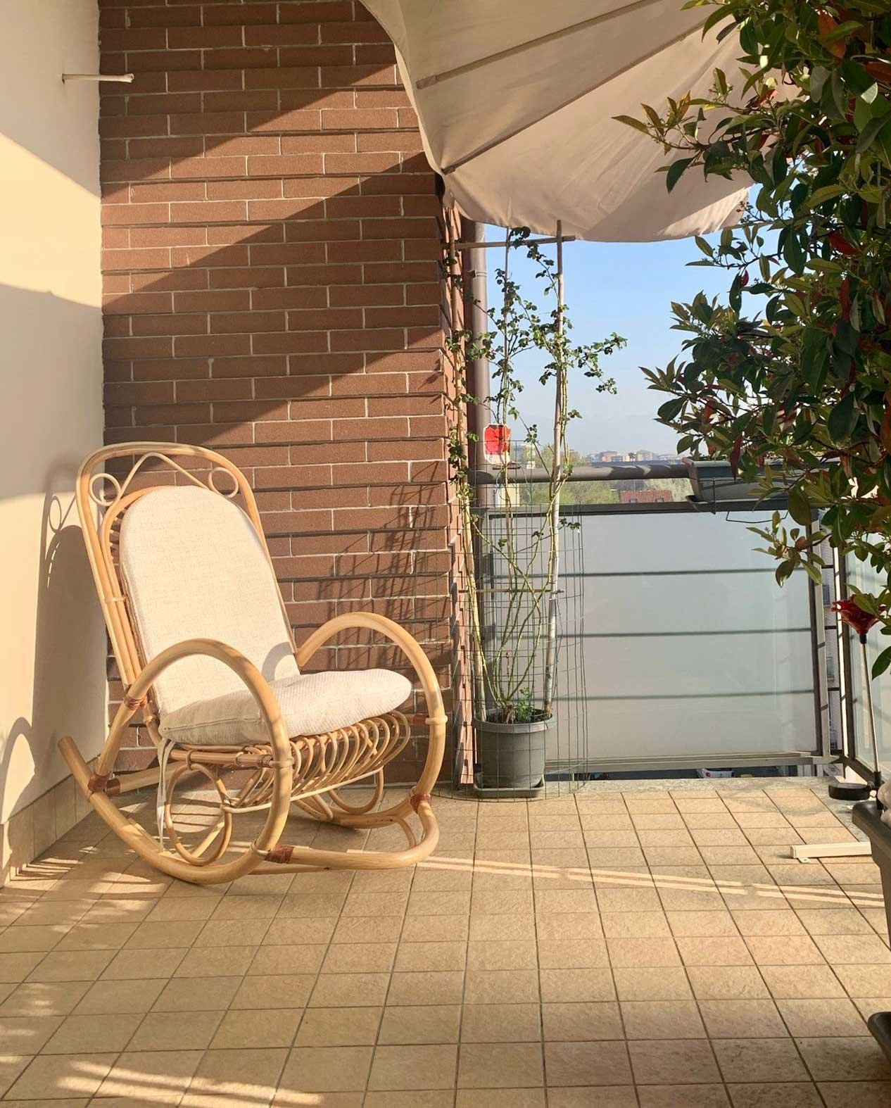 PoltronadondoloinrattancolorataG jollygiuncoeu wicker rocking chair on a sunlit balcony beside a brick wall with a potted plant and a white umbrella nearby