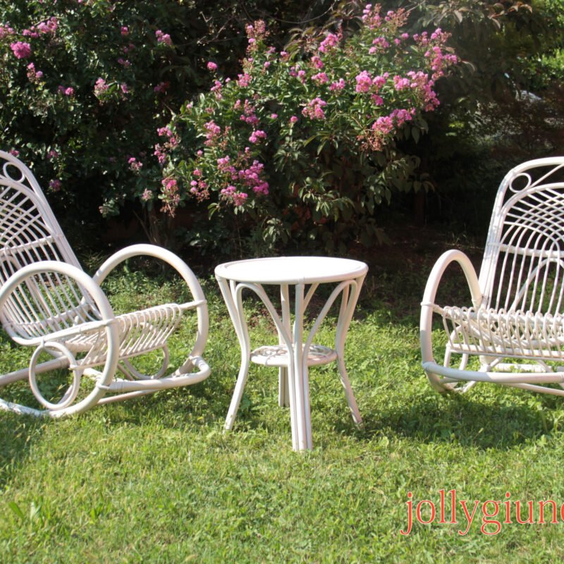 White metal outdoor table and chairs sit on a grassy lawn beside pink flowering bushes in a sunny garden.