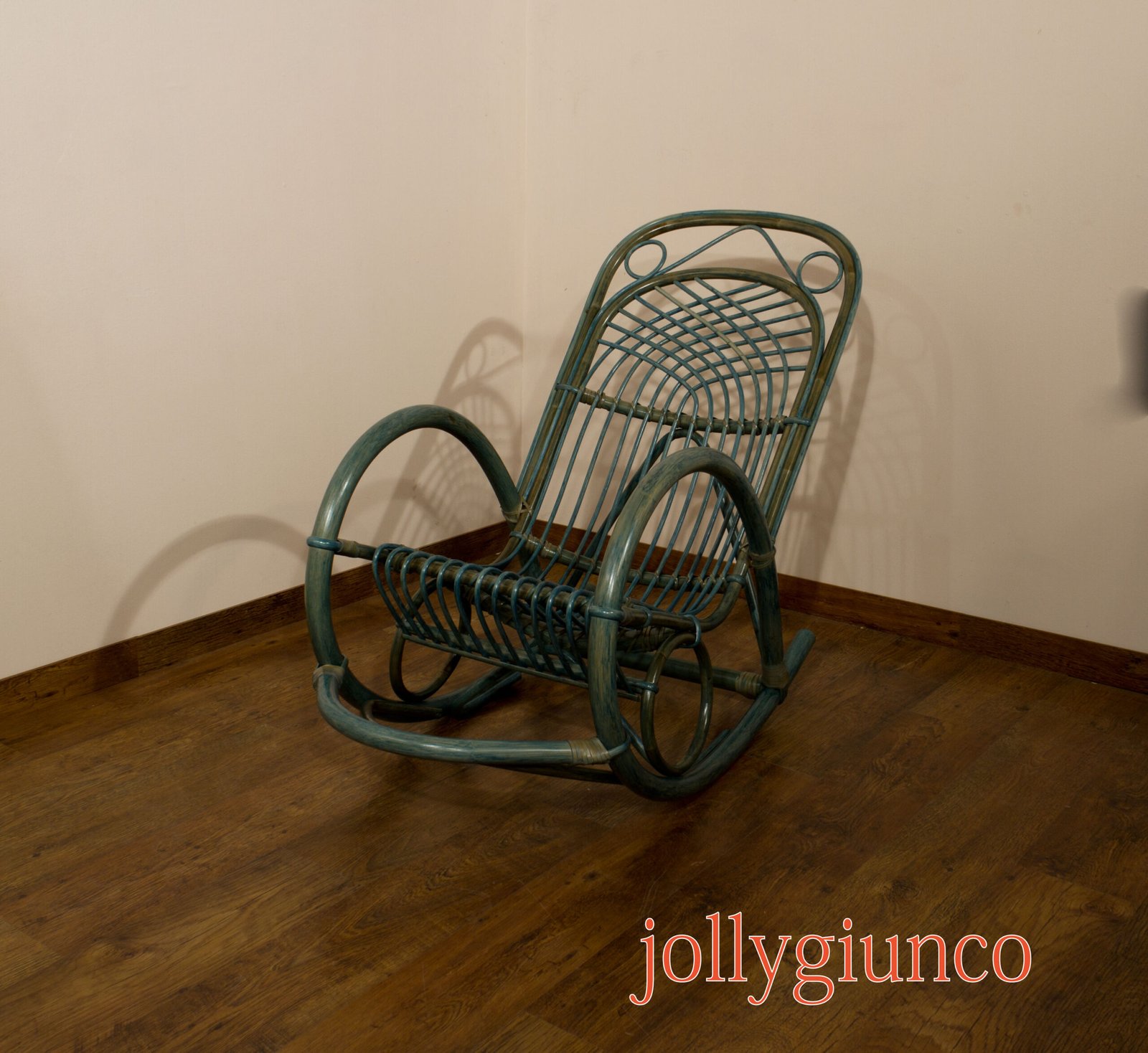 jollygiuncopoltrone8g jollygiuncoeu teal wicker rocking chair leaning to the right against a beige wall with a wooden floor visible beneath it