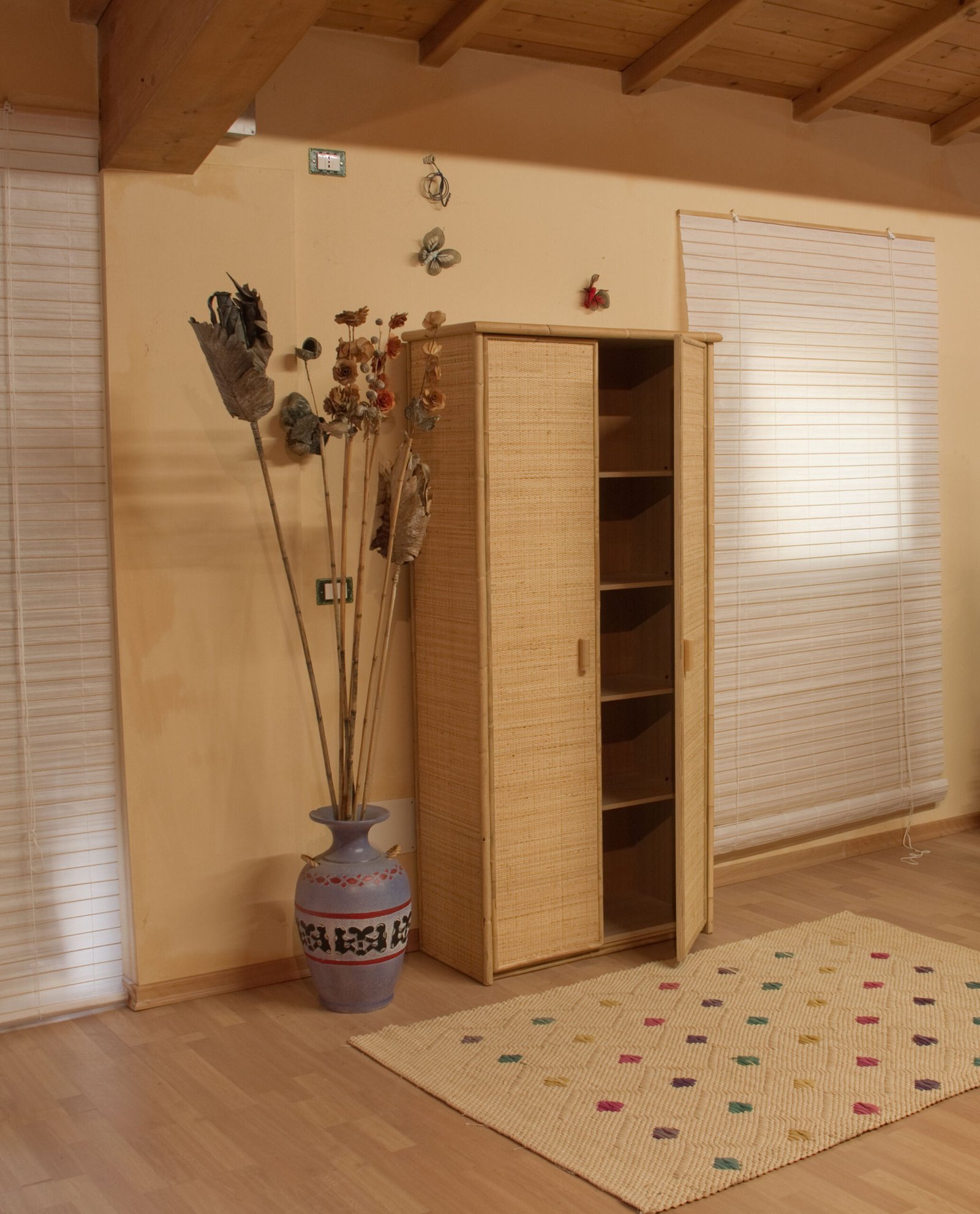 Colonna Cristina in giunco 2 porte jollygiuncoeu corner of a room with a tall vase of dried flowers beside a woven cabinet and beige walls with wooden ceiling beams visible above