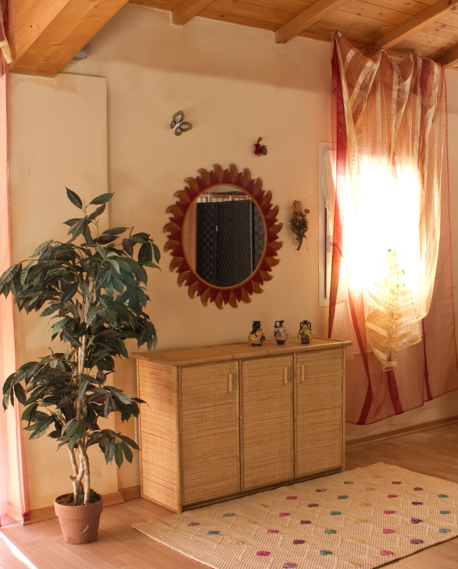 Mobile Patrizia in rattan 3 ante jollygiuncoeu corner of a living room with a tall potted plant a woven wicker cabinet and a sunburst round mirror on the wall beneath striped sheer curtains
