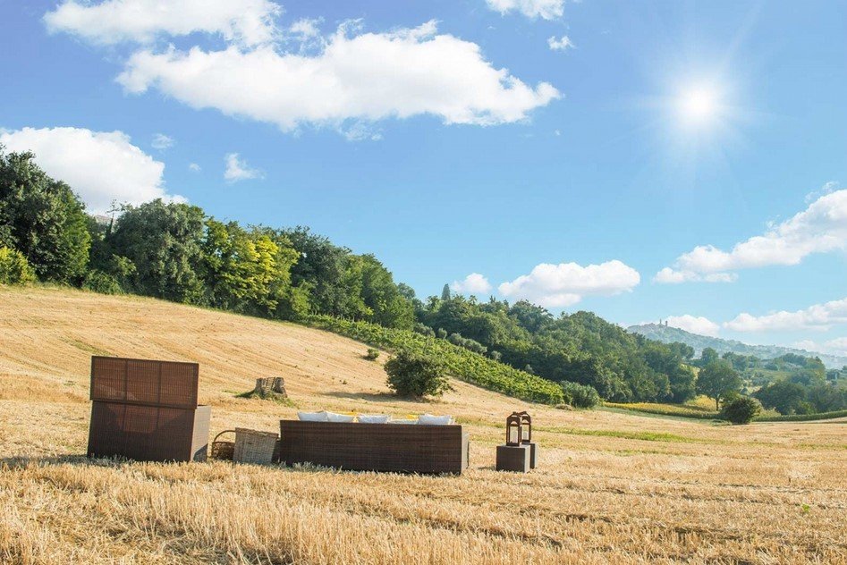 Cushion box baule Polyrattan4 jollygiuncoeu outdoor seating arranged in a sunlit golden field with a hillside and trees beyond under a bright blue sky