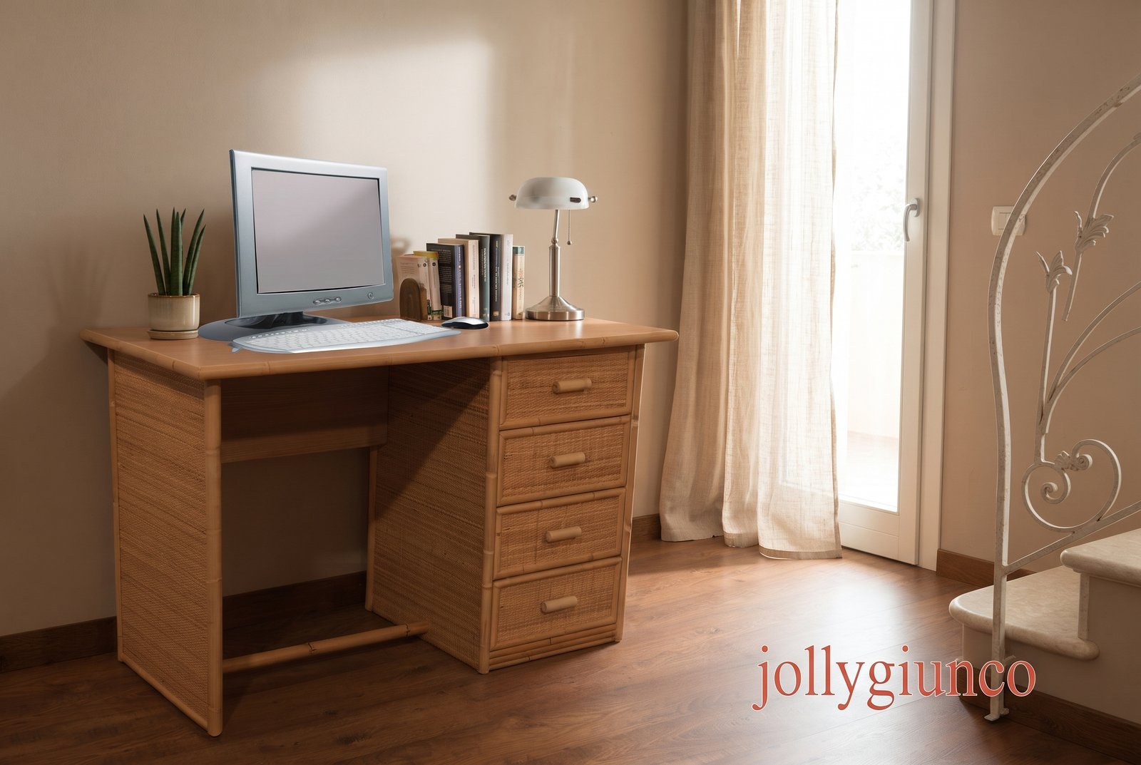 Scrivania salerno 4 Cassetti in Giunco jollygiuncoeu wooden desk with a small computer monitor white keyboard and mouse a row of books a metal desk lamp and a potted plant by a window with sheer curtains