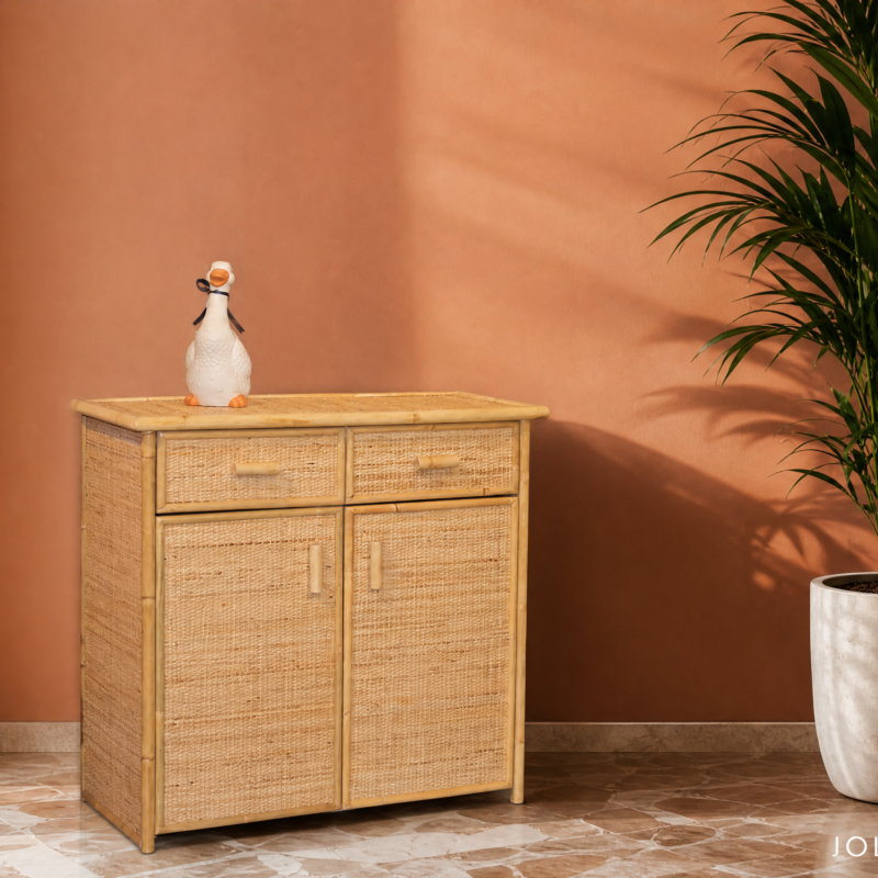 A woven wooden cabinet against a pink wall with a small white ceramic duck on top, and a large potted palm to the right; sheer curtain on the left and a marble-tiled floor visible at the base.