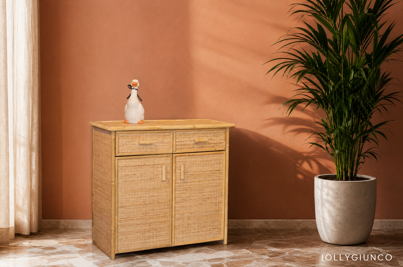 Scarpiera in Rattan 2 Porte e 1 Cassetto1 jollygiuncoeu a woven wooden cabinet against a pink wall with a small white ceramic duck on top and a large potted palm to the right sheer curtain on the left and a marble tiled floor visible at the base